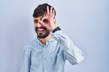 Young hispanic man with beard standing over blue background doing ok gesture with hand smiling, eye looking through fingers with happy face.