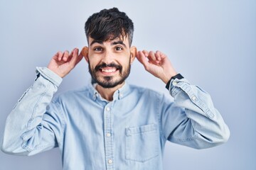 Young hispanic man with beard standing over blue background smiling pulling ears with fingers, funny gesture. audition problem