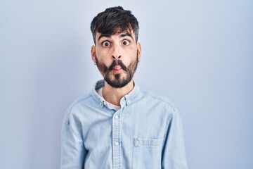 Young hispanic man with beard standing over blue background making fish face with lips, crazy and comical gesture. funny expression.