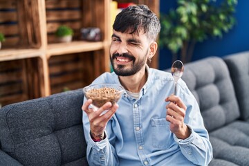 Young hispanic man with beard eating healthy whole grain cereals winking looking at the camera with sexy expression, cheerful and happy face.