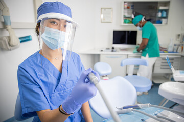 A female dentist in the clinic is holding the dental drill while her colleague stands behind.