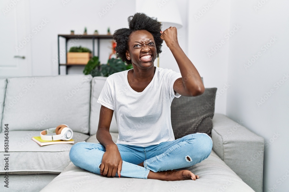 Wall mural african young woman sitting on the sofa at home angry and mad raising fist frustrated and furious wh