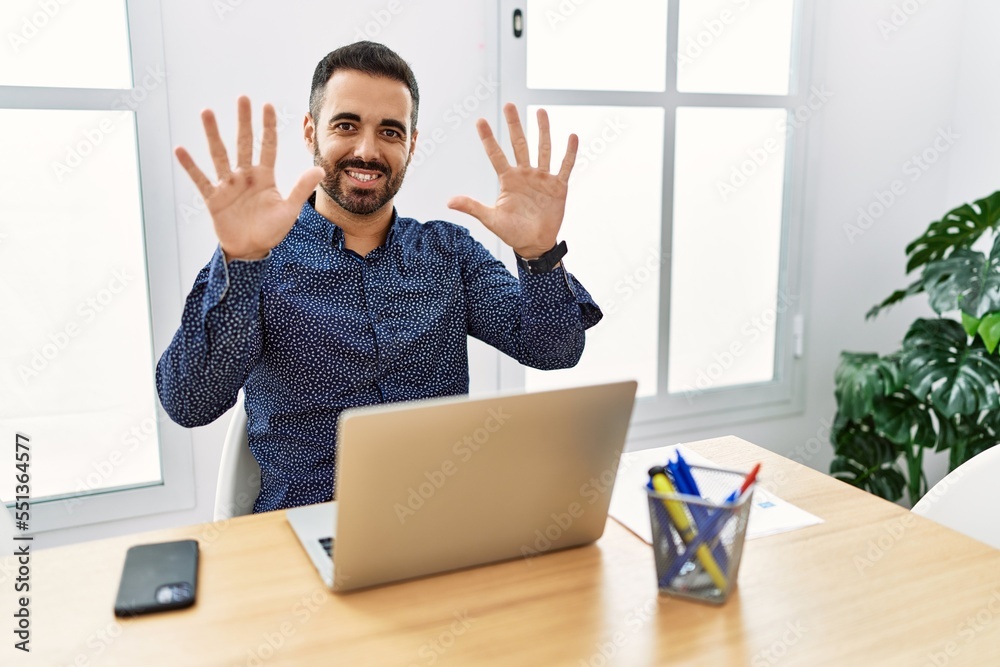 Sticker Young hispanic man with beard working at the office with laptop showing and pointing up with fingers number ten while smiling confident and happy.