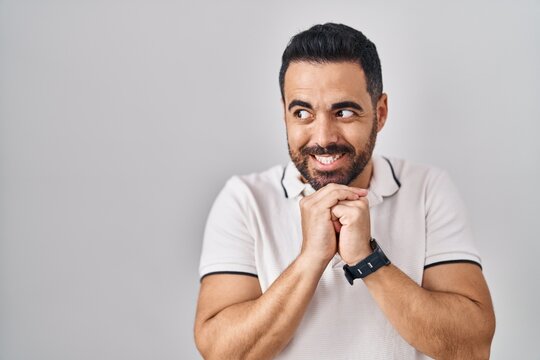Young hispanic man with beard wearing casual clothes over white background laughing nervous and excited with hands on chin looking to the side