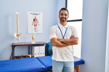 Young hispanic man physiotherapist standing with arms crossed gesture at rehab clinic