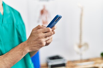 Young hispanic man wearing physiotherapist uniform using smartphone at physiotherapist clinic