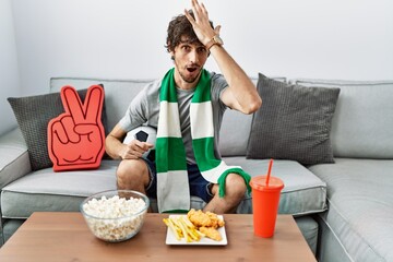 Young hispanic man football hooligan cheering game at home surprised with hand on head for mistake,...