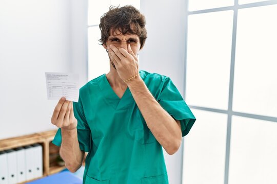 Young Hispanic Doctor Man Holding Covid Record Card Smelling Something Stinky And Disgusting, Intolerable Smell, Holding Breath With Fingers On Nose. Bad Smell