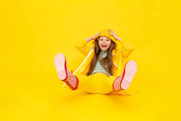 A child in a raincoat, sitting with his legs up in red boots. A beautiful little girl is smiling and sitting on a yellow isolated background. Rainy spring, autumn weather.