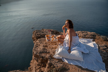 Woman covered with a blanket of bed relaxing and watching the seascape at sunrise. She holds a cup of coffee in her hand in front of her is a table with fruits and croissants. Wanderlust and freedom