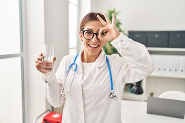 Young brunette doctor woman holding glass of water smiling happy doing ok sign with hand on eye looking through fingers