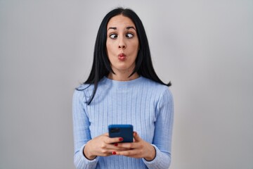 Hispanic woman using smartphone making fish face with mouth and squinting eyes, crazy and comical.