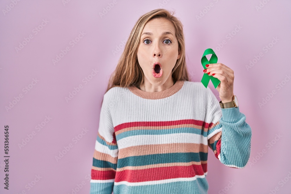 Poster young blonde woman holding support green ribbon scared and amazed with open mouth for surprise, disb