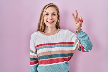 Young blonde woman standing over pink background showing and pointing up with fingers number two while smiling confident and happy.