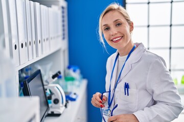 Young blonde woman scientist smiling confident holding glasses at laboratory