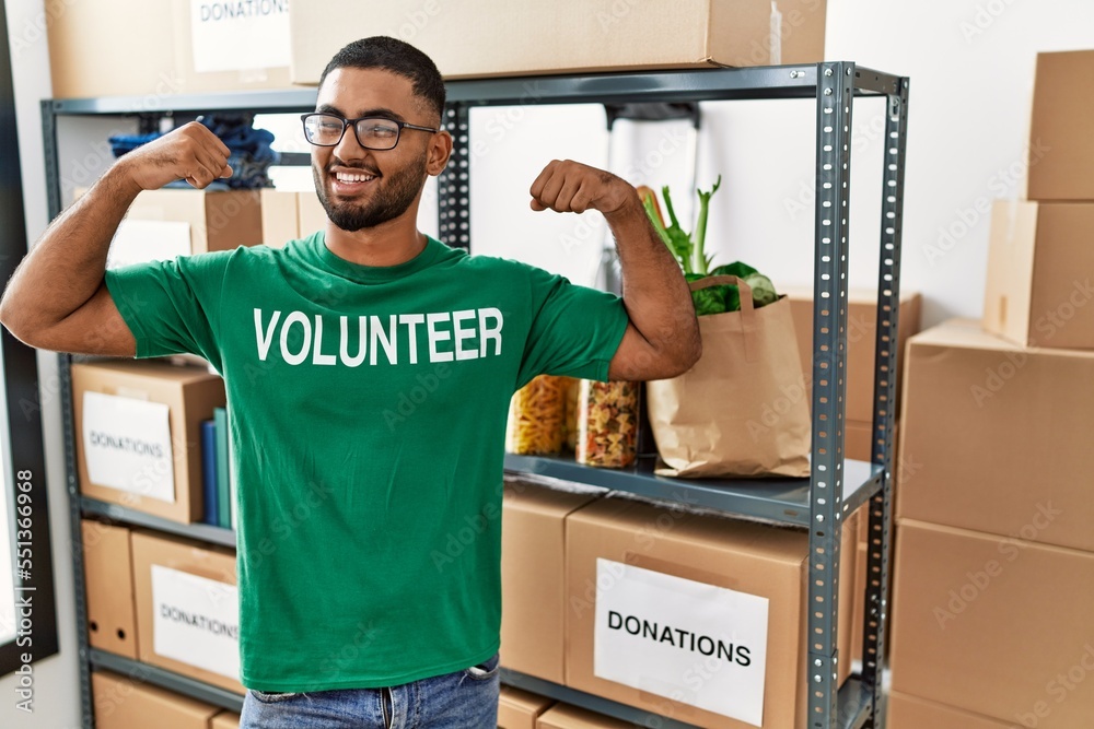 Sticker young indian man volunteer holding donations box showing arms muscles smiling proud. fitness concept