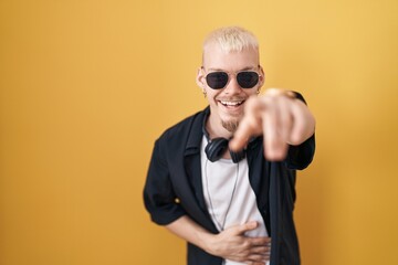 Fototapeta premium Young caucasian man wearing sunglasses standing over yellow background laughing at you, pointing finger to the camera with hand over body, shame expression