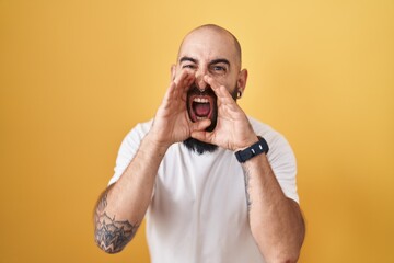 Young hispanic man with beard and tattoos standing over yellow background shouting angry out loud with hands over mouth