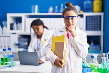 Mother and young daughter working at scientist laboratory serious face thinking about question with...