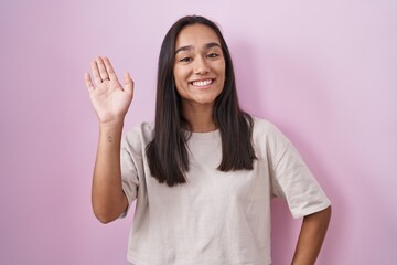 Young hispanic woman standing over pink background waiving saying hello happy and smiling, friendly...