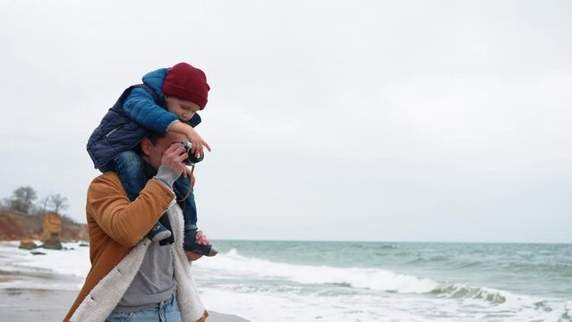 Brunet father takes photo on camera while his son on his neck on the beach 