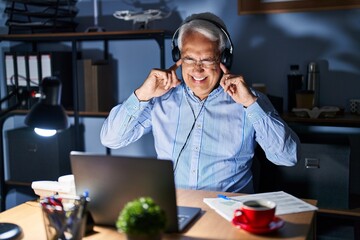 Hispanic senior man wearing call center agent headset at night covering ears with fingers with annoyed expression for the noise of loud music. deaf concept.