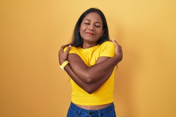 Young indian woman standing over yellow background hugging oneself happy and positive, smiling...