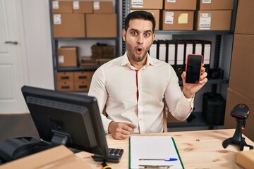 Fototapeta na wymiar Young hispanic man with beard working at small business ecommerce showing smartphone screen scared and amazed with open mouth for surprise, disbelief face