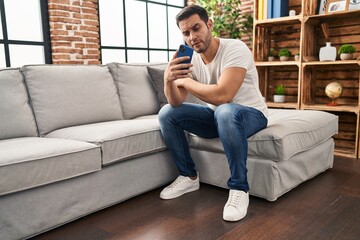 Fototapeta premium Young hispanic man using smartphone sitting on sofa at home