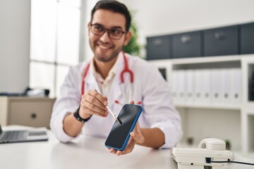 Young hispanic man wearing doctor uniform showing medical smartphone app at clinic
