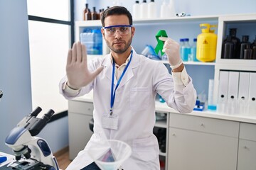 Young hispanic man with beard working at scientist laboratory holding green ribbon with open hand doing stop sign with serious and confident expression, defense gesture