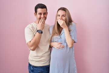 Young couple expecting a baby standing over pink background laughing and embarrassed giggle...