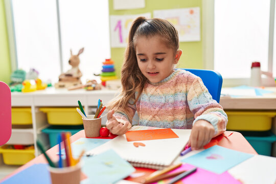 Adorable hispanic girl student sitting on table drawing on paper at kindergarten