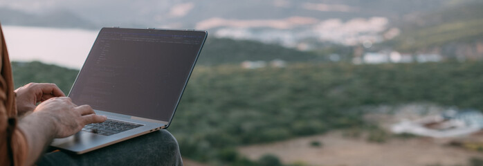 A man remotely works with a laptop on a mountain with a view of the mountains. Close-up of hands...