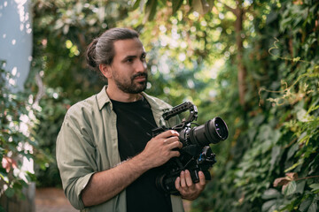 A young male cameraman with a professional movie camera in his hands in a green garden.