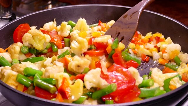 Cooking vegetable stew from frozen chopped vegetables. Tomatoes, asparagus and cabbage are steaming in a pan. Stir the dish with a kitchen spatula.