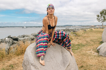 Pretty Asian woman with barefeet wearing colorful striped pants, a visor and black bikini top outside near water