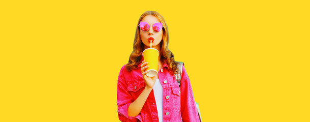 Portrait of stylish young woman drinking juice wearing pink jacket, sunglasses on yellow background