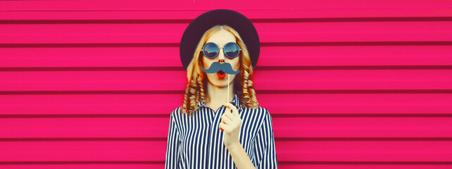 Portrait of funny cheerful young woman having fun showing mustache on stick on pink background