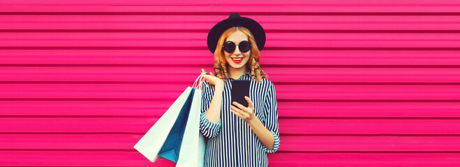 Portrait of happy smiling woman with shopping bags and smartphone wearing striped shirt, black round hat on pink background