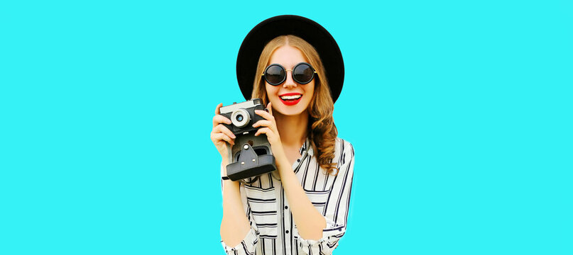 Portrait of happy smiling young woman photographer taking picture on film camera wearing black round hat, striped shirt isolated on blue background