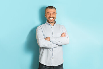 Male portrait. Happy middle aged caucasian man with folded arms smiling at camera, posing over blue studio background