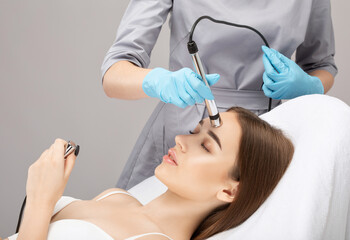 Naklejka premium A doctor cosmetologist makes a microcurrent facial therapy to a young woman with a device in a beauty wellness salon.Cosmetology and professional skin care.