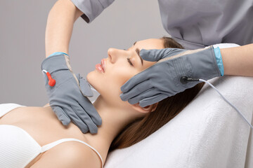A cosmetologist doctor makes a microcurrent massage procedure on the face and neck of a young...