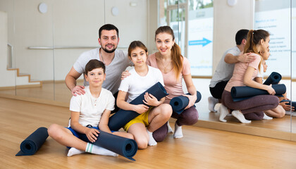 Fototapeta na wymiar Cheerful friendly sporty young parents with teen daughter and son posing with gymnastic mats before exercising in yoga class