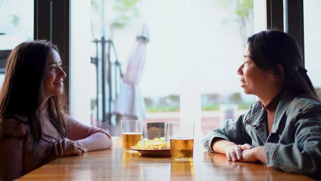Two girls sitting at table of a bar hanging out while they talk with each other with beers