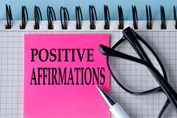 POSITIVE AFFIRMATIONS - words on a pink piece of paper on a white notebook with glasses and pen
