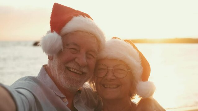 Two old happy seniors wearing christmas hats at the beach taking a selfie of them smiling and having fun with the sunset at the background at evening. Cute couple of old persons looking at the camera 