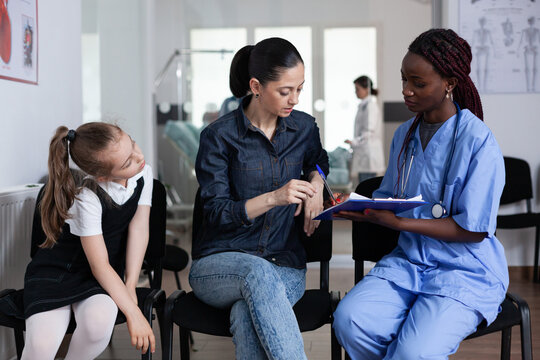 Young Woman Signing Little Girl Discharge Papers In Children Hospital Waiting Room. Mother Filling Out Daughter File With African American Pediatrician At Medical Clinic Lobby.