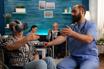 Elderly woman uses a virtual reality headset on a wheelchair in a nursing home to explore augmented reality. Male nurse in common room of skilled care facility helping patient to have some fun.